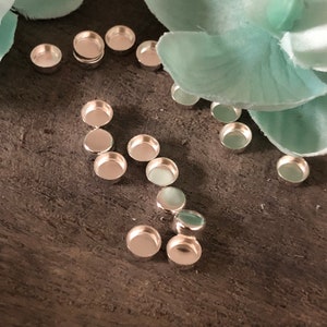 Puede incluir: Un conjunto de bandejas redondas de metal plateado para la elaboraci&oacute;n de joyas. Las bandejas est&aacute;n dispuestas sobre una superficie de madera con una flor azul claro.