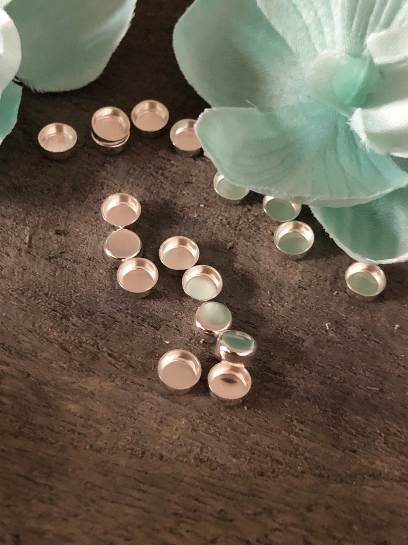 Puede incluir: Un conjunto de bandejas redondas de metal plateado para la elaboraci&oacute;n de joyas. Las bandejas est&aacute;n dispuestas sobre una superficie de madera con una flor azul claro.