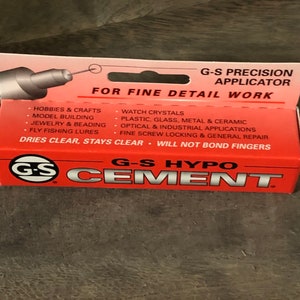 Op de afbeelding: Een rood en wit pakket G-S Hypo Cement met een precisie-applicator voor fijn detailwerk. De verpakking vermeldt toepassingen voor hobby's en ambachten, modelbouw, sieraden en kralen, vliegvissen, horlogekristallen, kunststof, glas, metaal en keramiek, optische en industri&euml;le toepassingen, fijne schroefvergrendeling en algemene reparatie. De verpakking geeft aan dat de lijm helder droogt, helder blijft en niet aan de vingers kleeft.