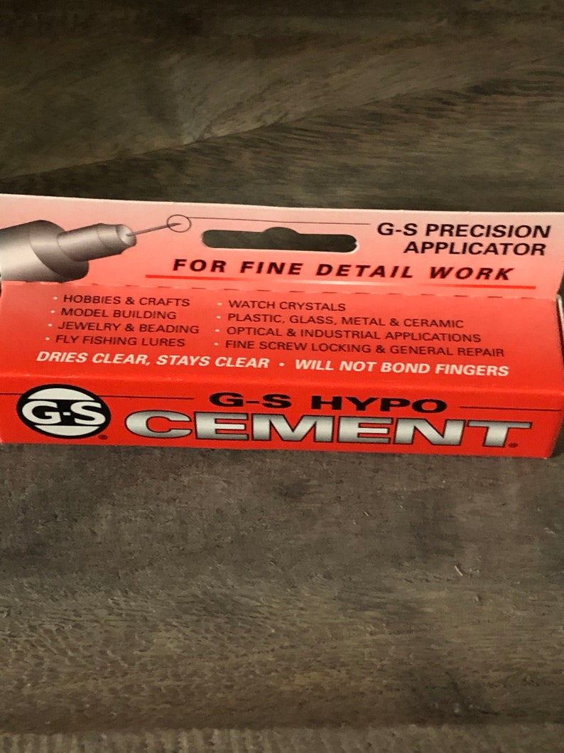 Op de afbeelding: Een rood en wit pakket G-S Hypo Cement met een precisie-applicator voor fijn detailwerk. De verpakking vermeldt toepassingen voor hobby's en ambachten, modelbouw, sieraden en kralen, vliegvissen, horlogekristallen, kunststof, glas, metaal en keramiek, optische en industri&euml;le toepassingen, fijne schroefvergrendeling en algemene reparatie. De verpakking geeft aan dat de lijm helder droogt, helder blijft en niet aan de vingers kleeft.
