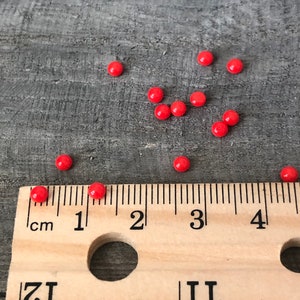 May include: A close-up of a wooden ruler with red beads scattered around it. The ruler is marked in inches and centimeters. The beads are about 1/4 inch in diameter.