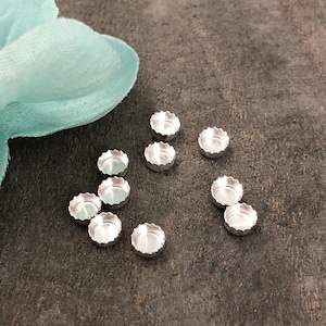 May include: A collection of small, silver-colored metal components with a scalloped edge, scattered on a dark, textured surface. A soft, light blue fabric is visible in the upper left corner. These components appear to be small metal caps.
