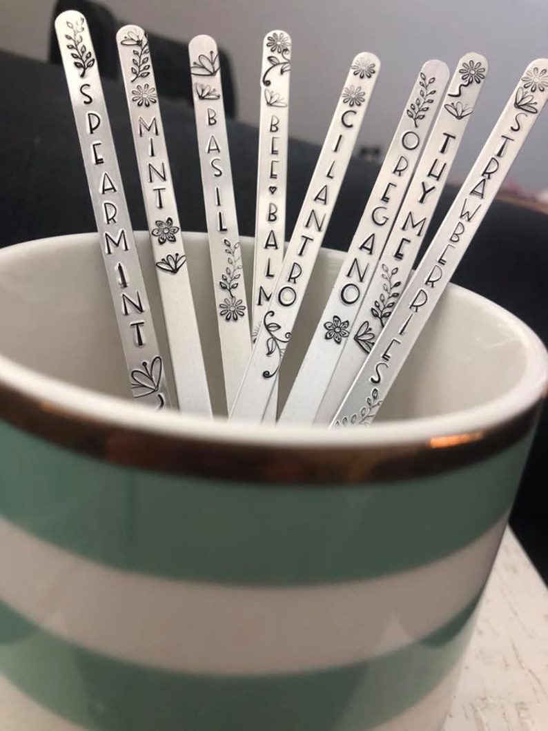 May include: Silver metal plant markers with engraved names of herbs, including Spearmint, Mint, Basil, Bee Balm, Cilantro, Oregano, Thyme, Strawberry. The markers are in a striped mug.