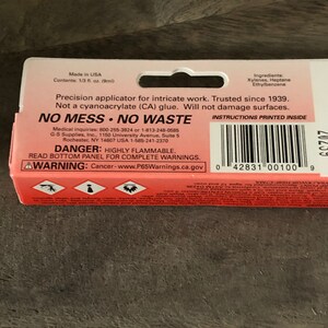 Op de afbeelding: Rood en wit etiket met de tekst "NO MESS NO WASTE" en "DANGER: HIGHLY FLAMMABLE." Het etiket is voor een precisie-applicator voor ingewikkeld werk. Het etiket bevat ook instructies die binnenin zijn gedrukt en een waarschuwing voor kanker.
