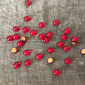 May include: A scattering of red glass beads on a gray wooden surface. The beads are small and round, and they are arranged in a random pattern.