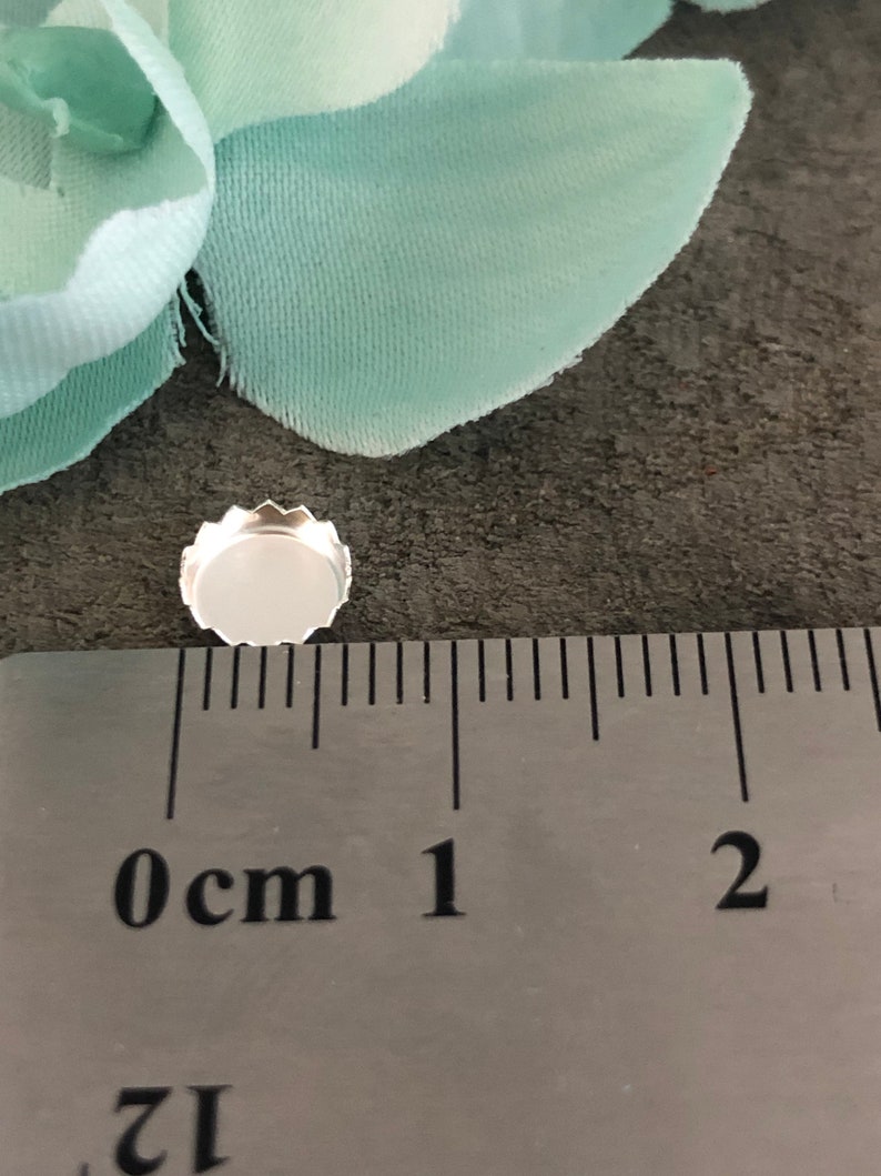 May include: A silver-coloured metal earring setting with a 0.5 cm diameter, measured against a ruler. The setting is round with a scalloped edge.