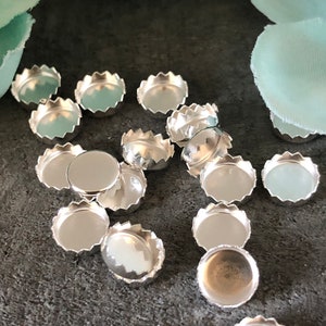 May include: A collection of silver-toned metal bezel settings for jewelry making. The bezels have a scalloped edge and are scattered on a gray surface.