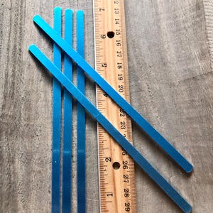May include: Four blue metal strips, each approximately 9 inches long, are arranged on a wooden surface next to a ruler. The ruler is marked in inches and shows the length of the strips.