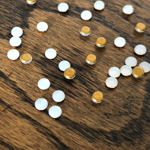May include: A collection of small, round, white and gold embellishments. The embellishments are scattered on a wooden surface.