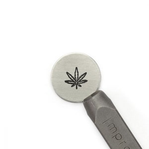 May include: A silver metal stamping blank with a black cannabis leaf design. A metal stamping tool is positioned next to the blank. The background is white.