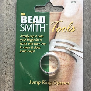 May include: A green and white packaging card for a jump ring opener tool. The card features the text "the BEAD SMITH Tools" and "Jump Ring Opener". The tool is a silver ring that can be worn on a finger to open and close jump rings.