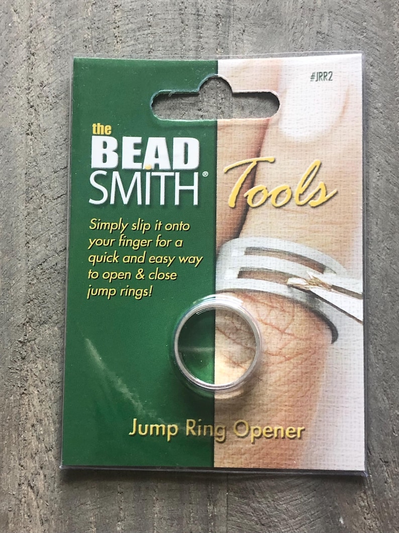 May include: A green and white packaging card for a jump ring opener tool. The card features the text "the BEAD SMITH Tools" and "Jump Ring Opener". The tool is a silver ring that can be worn on a finger to open and close jump rings.