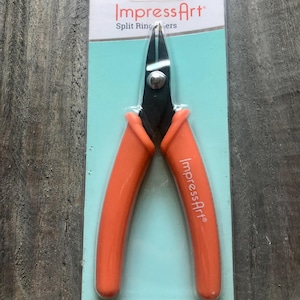 May include: Orange handled pliers with black metal jaws, packaged in a clear plastic clamshell with a blue and white background. The packaging reads "ImpressArt Split Ring Pliers".
