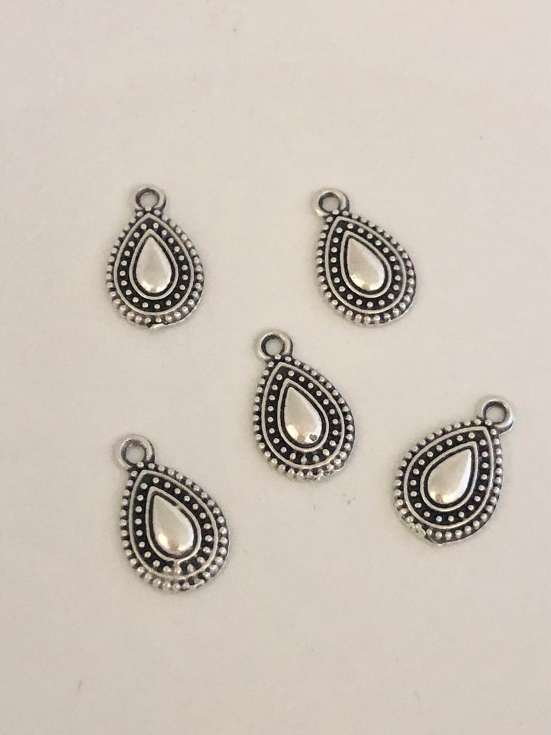 May include: Five silver-colored teardrop charms with a textured border of small dots. Each charm has a loop at the top for attaching to jewelry. The teardrop shape has a smooth, reflective center.