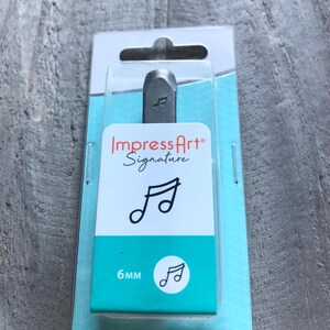 May include: A silver metal stamp with a music note design. The stamp is packaged in a clear plastic blister pack with a blue and white background. The packaging includes the brand name "ImpressArt Signature" and the size of the stamp, "6mm".