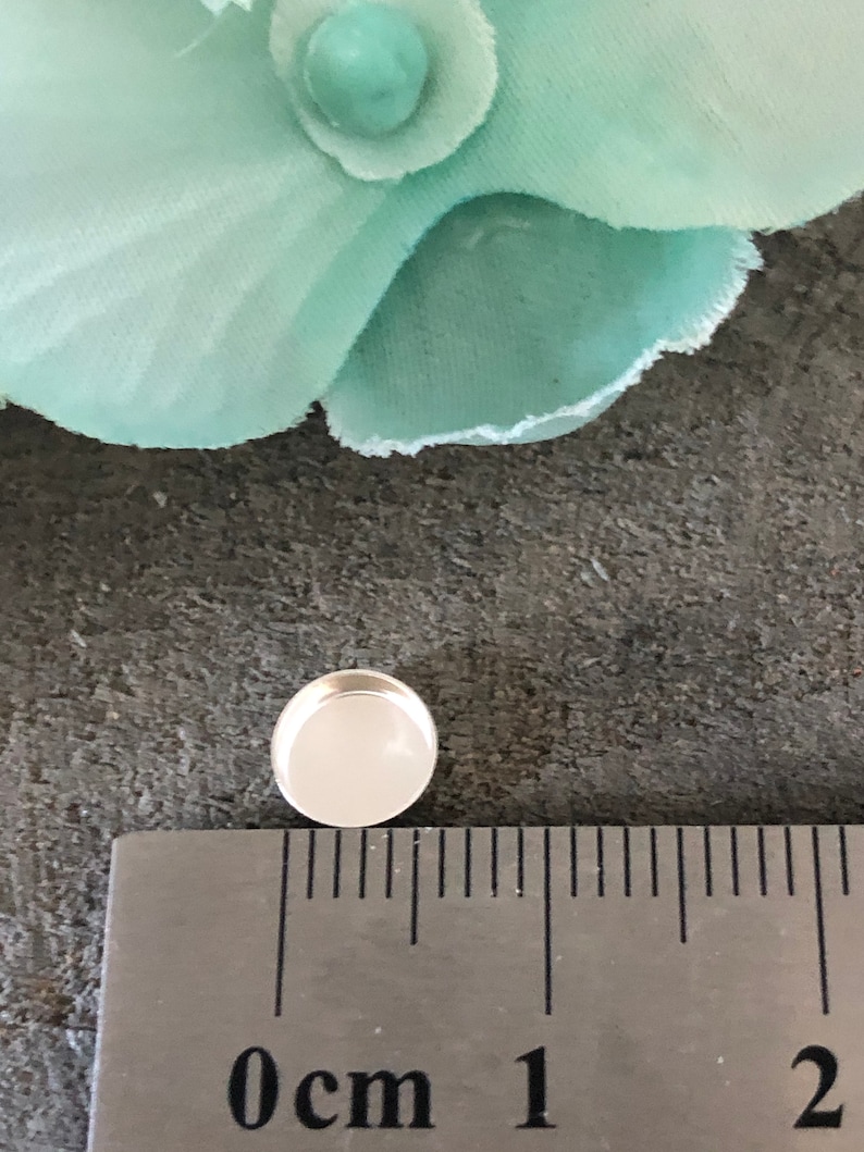 May include: A silver-colored, round, flat-backed cabochon with a white center, measuring approximately 1 cm in diameter.