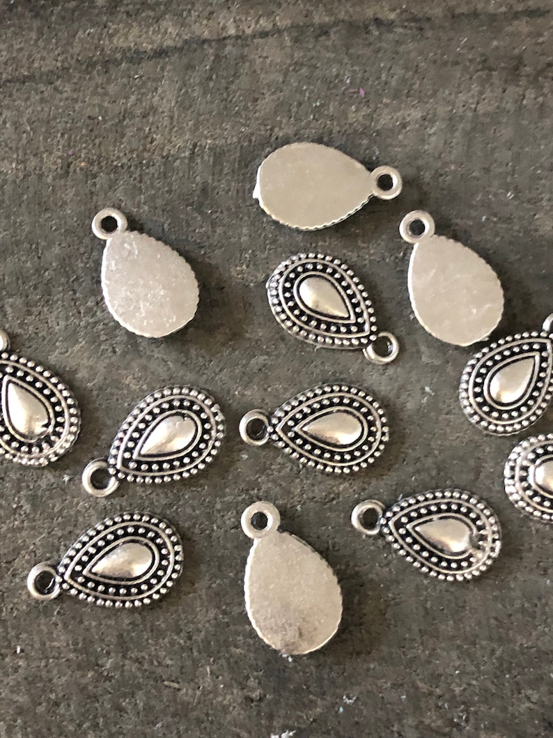 May include: Silver-toned teardrop-shaped charms with a small loop at the top for stringing. Some charms have a detailed beaded border around a smooth, teardrop-shaped center. The charms are scattered on a dark gray surface.