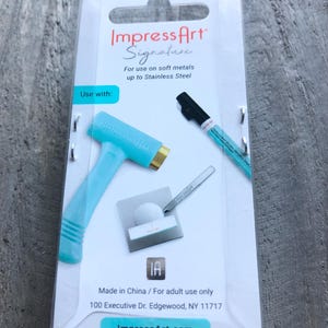 May include: A turquoise blue metal stamping tool with a gold head, a black marker pen, and a small metal stylus. The packaging includes the text "Impress Art Signature" and "For use on soft metals up to Stainless Steel".