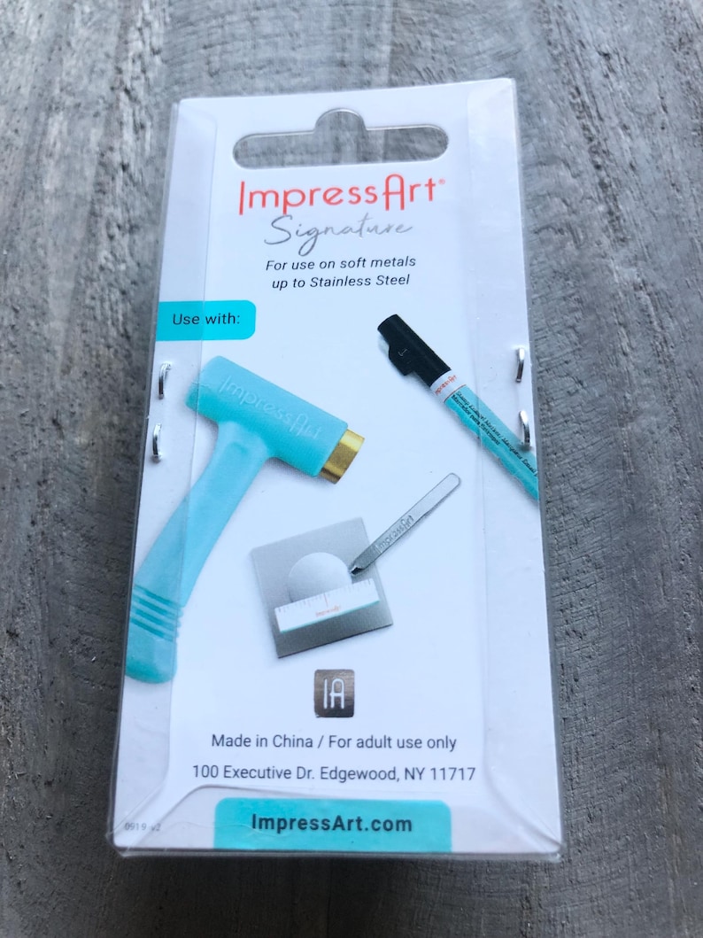 May include: A turquoise blue metal stamping tool with a gold head, a black marker pen, and a small metal stylus. The packaging includes the text "Impress Art Signature" and "For use on soft metals up to Stainless Steel".