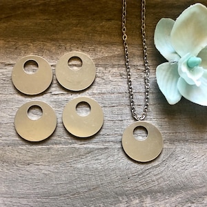 May include: Four silver metal circles with a hole in the center, and a silver chain necklace with a similar circle pendant. The circles are on a wooden surface.