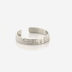May include: A silver toe ring with a textured surface. The ring is open and adjustable.