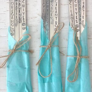 May include: A set of nine metal plant markers with different herb names, including oregano, basil, sage, chives, cilantro, rosemary, and thyme. The markers are wrapped in light blue tissue paper and tied with twine.