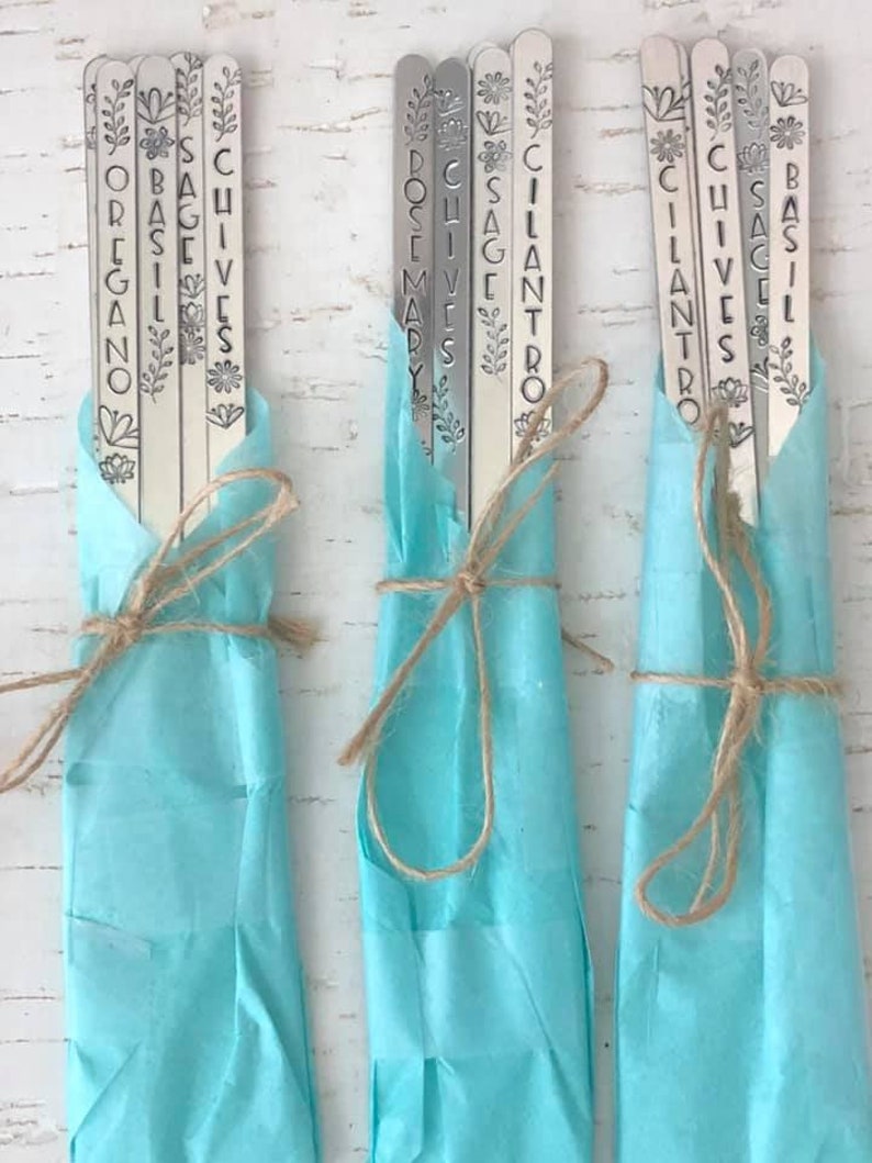 May include: A set of nine metal plant markers with different herb names, including oregano, basil, sage, chives, cilantro, rosemary, and thyme. The markers are wrapped in light blue tissue paper and tied with twine.