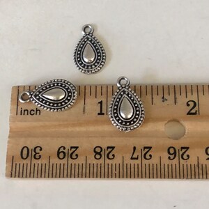 May include: Three silver-colored teardrop charms with a textured border and a loop at the top. The charms are next to a wooden ruler, with the largest charm measuring approximately 1 inch in length.