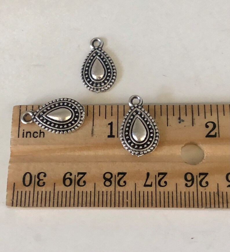 May include: Three silver-colored teardrop charms with a textured border and a loop at the top. The charms are next to a wooden ruler, with the largest charm measuring approximately 1 inch in length.
