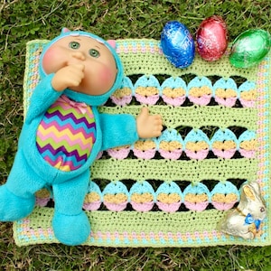 May include: A crocheted baby blanket with a pastel green, pink, and blue color scheme. The blanket features a repeating pattern of crocheted eggs. A Cabbage Patch Kid doll wearing a blue and pink outfit is lying on the blanket. A chocolate bunny and three foil-wrapped chocolate eggs are also on the blanket.