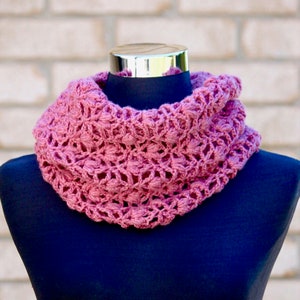 May include: A pink crocheted infinity scarf with a delicate openwork pattern. The scarf is displayed on a black mannequin.