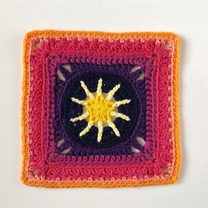 May include: A crocheted square with a colourful gradient border in shades of orange, pink, and purple. The centre features a yellow starburst design.