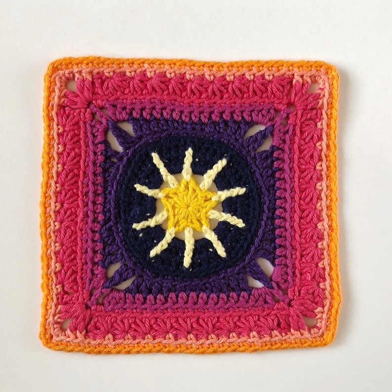May include: A crocheted square with a colourful gradient border in shades of orange, pink, and purple. The centre features a yellow starburst design.