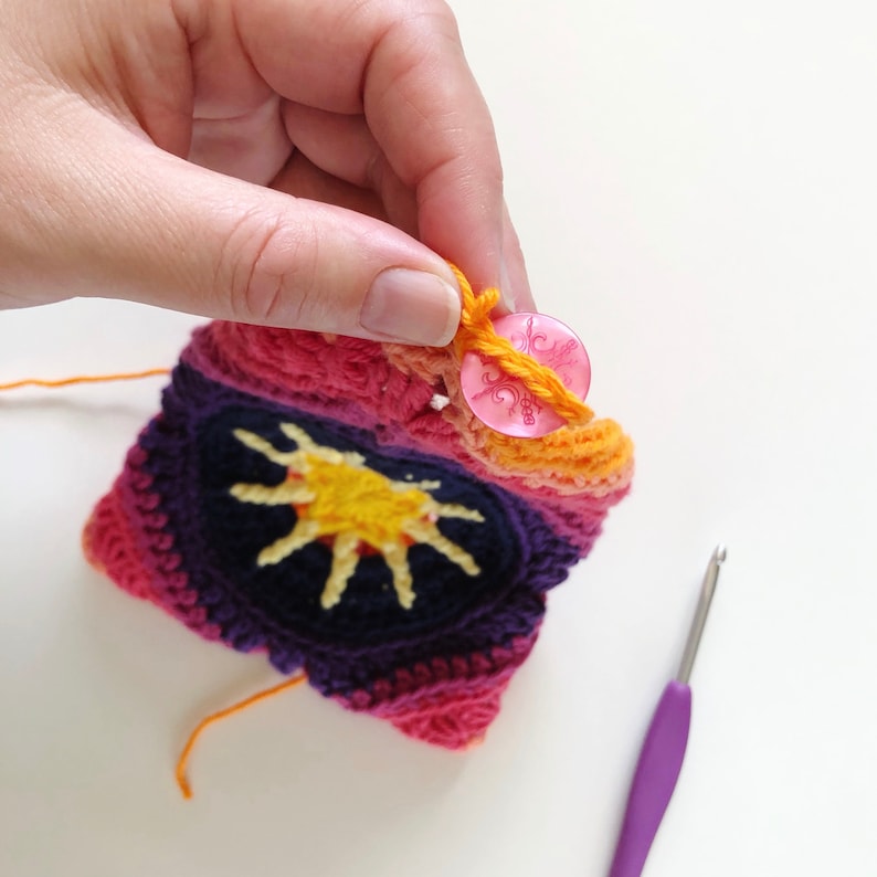 May include: A crocheted sunburst design with a pink button and orange yarn. The crocheted piece is in shades of pink, purple, and orange.