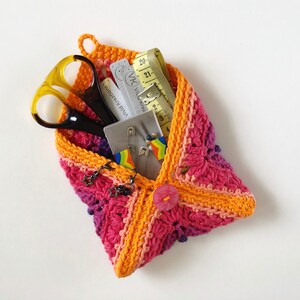May include: A crocheted pouch in shades of pink, orange, and purple, filled with sewing supplies, including scissors, measuring tape, safety pins, and a small card with the text "Viva Kreations".