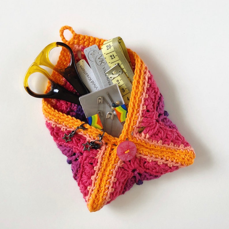 May include: A crocheted pouch in shades of pink, orange, and purple, filled with sewing supplies, including scissors, measuring tape, safety pins, and a small card with the text "Viva Kreations".