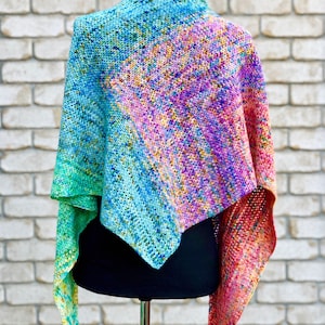 Puede incluir: Un chal de ganchillo colorido con un degradado de azul, verde, rosa y naranja. El chal está drapeado sobre un maniquí negro.