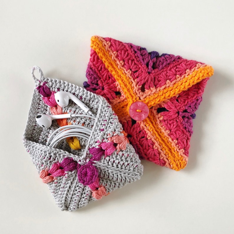 May include: Two crocheted pouches, one grey with pink and orange accents and one with a gradient of pink, orange, and yellow. The grey pouch has a button closure and is filled with white earbuds.