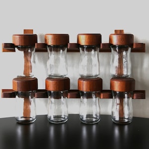 Puede incluir: Un juego de ocho tarros de especias de vidrio con tapas de madera, montados en un estante de madera. Los tarros son transparentes y las tapas son de color marrón oscuro.