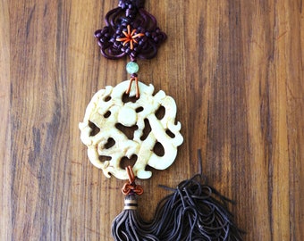Op de afbeelding: Een witte jade hanger met een gedetailleerd gesneden ontwerp hangt aan een bruin koord met een kwast. Het koord is versierd met een bruin en paarse knoop.