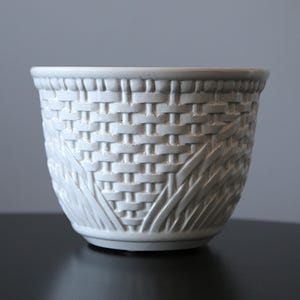 May include: A white ceramic plant pot with a woven basket design. The pot has a round shape and a textured surface, with a detailed pattern of interwoven lines. Suitable for indoor or outdoor use.