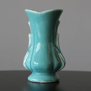 Può includere: Un vaso in ceramica turchese con un design scanalato. Il vaso ha una parte superiore svasata e una base a piedistallo. La finitura lucida riflette la luce, evidenziando le curve e i dettagli di questo pezzo decorativo vintage. È un oggetto decorativo.