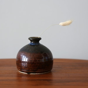 May include: A small, round ceramic vase with a dark brown and blue glaze. The vase holds a single, dried, beige-colored stem. The vase is displayed on a wooden surface, showcasing its handcrafted nature and minimalist design.