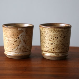 May include: Two handmade ceramic cups with a speckled, tan glaze. The cups have a cylindrical shape with a slightly flared rim and a dark brown band around the top. The base is a darker brown color.