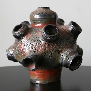 Puede incluir: Un jarrón de cerámica con una superficie texturizada y moteada en tonos de negro, gris y rojo. El jarrón tiene una base redonda y múltiples aberturas alrededor de la parte superior, que se asemejan a un capullo de flor.