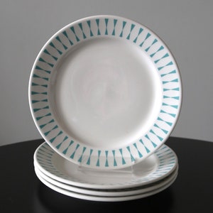 Puede incluir: Cuatro platos de cerámica blanca con un patrón geométrico turquesa en el borde. Apilados sobre una superficie negra contra un fondo gris. Diseño sencillo y elegante.