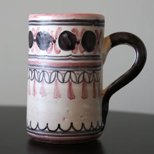 May include: A white ceramic mug with a brown handle. The mug has a pink and black geometric pattern.