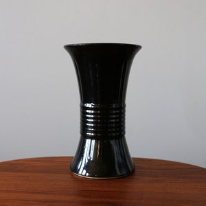 Puede incluir: Un jarrón de cerámica negro con la parte superior acampanada y la base estrecha. El jarrón tiene una banda de líneas elevadas alrededor del centro.