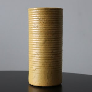 Puede incluir: Un jarrón cilíndrico de color amarillo mostaza con detalles horizontales acanalados. El jarrón de cerámica tiene una superficie ligeramente texturizada y un diseño simple y minimalista. El jarrón es adecuado para exhibir flores o como pieza decorativa.