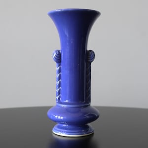 Könnte beinhalten: Eine kobaltblaue Keramikvase mit ausgestelltem Oberteil und bauchigem Boden. Die Vase hat dekorative seilartige Details an den Seiten und zwei kleine, kreisförmige Griffe. Die Vase ist etwa 20 cm hoch.